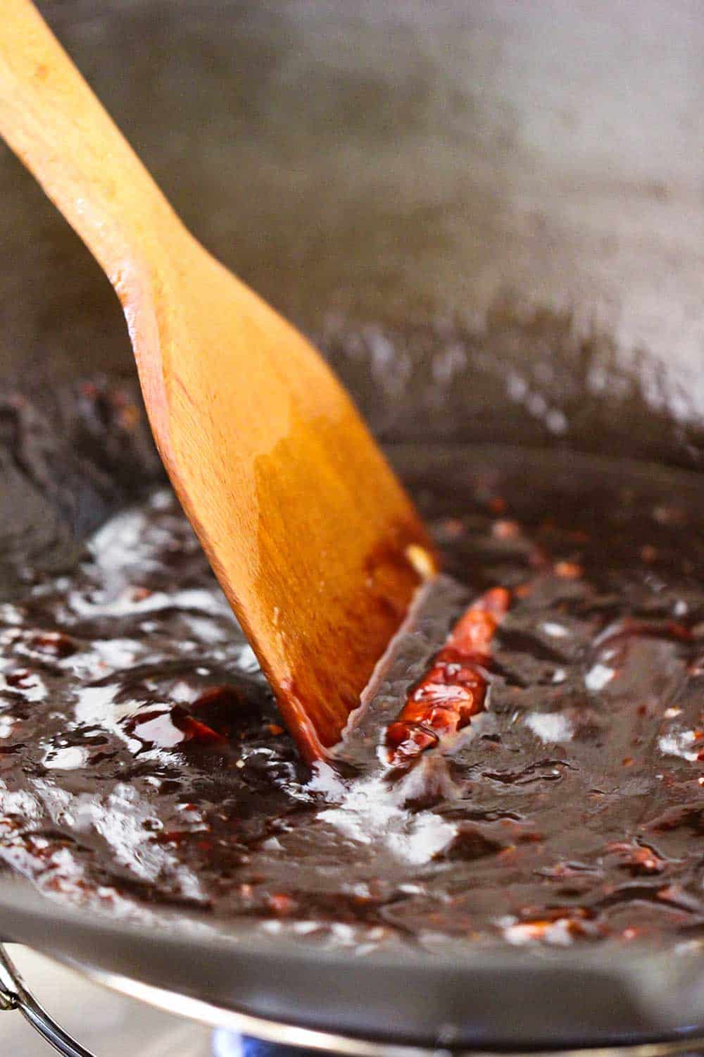 A wooden spoon stirring a thick brown sauce in a large wok. 