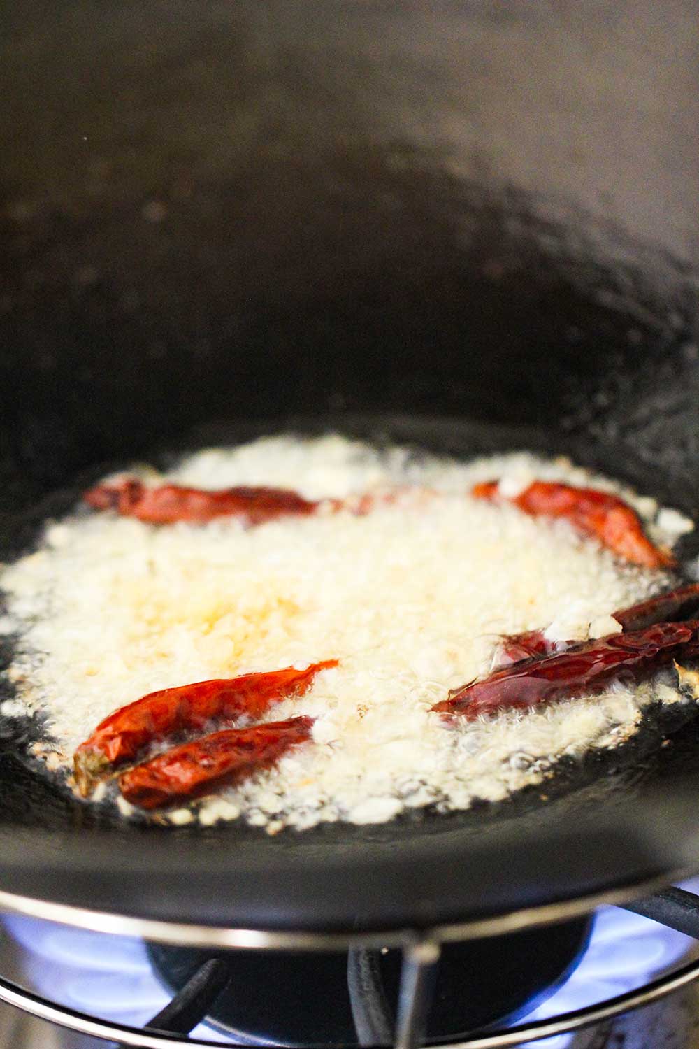 A wok over a flame with chopped ginger, garlic, and red chili peppers cooking in hot oil. 