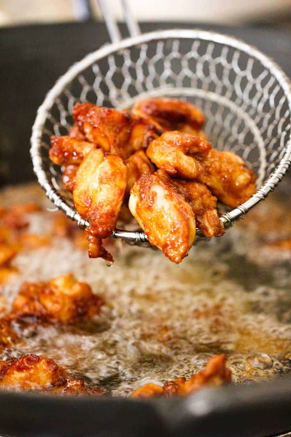 A slotted metal spoon holding pieces of fried chicken over a wok filled with hot oil. 