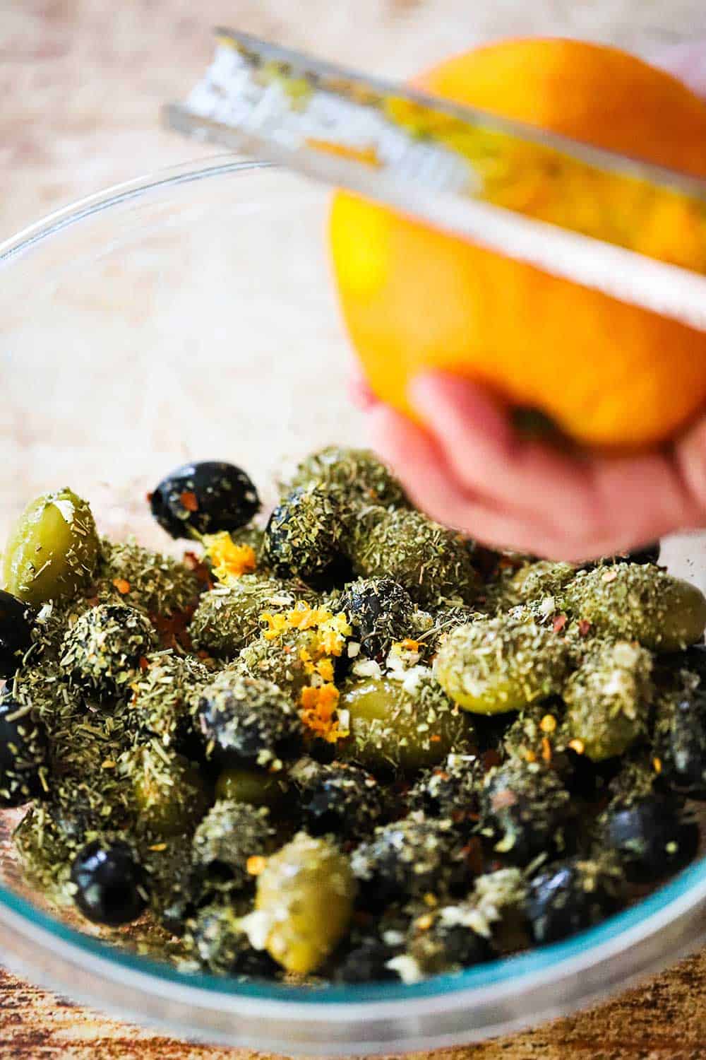 A person using a citrus zester to collect zest from an orange over a bowl of black and green olives topped with dried herbs. 