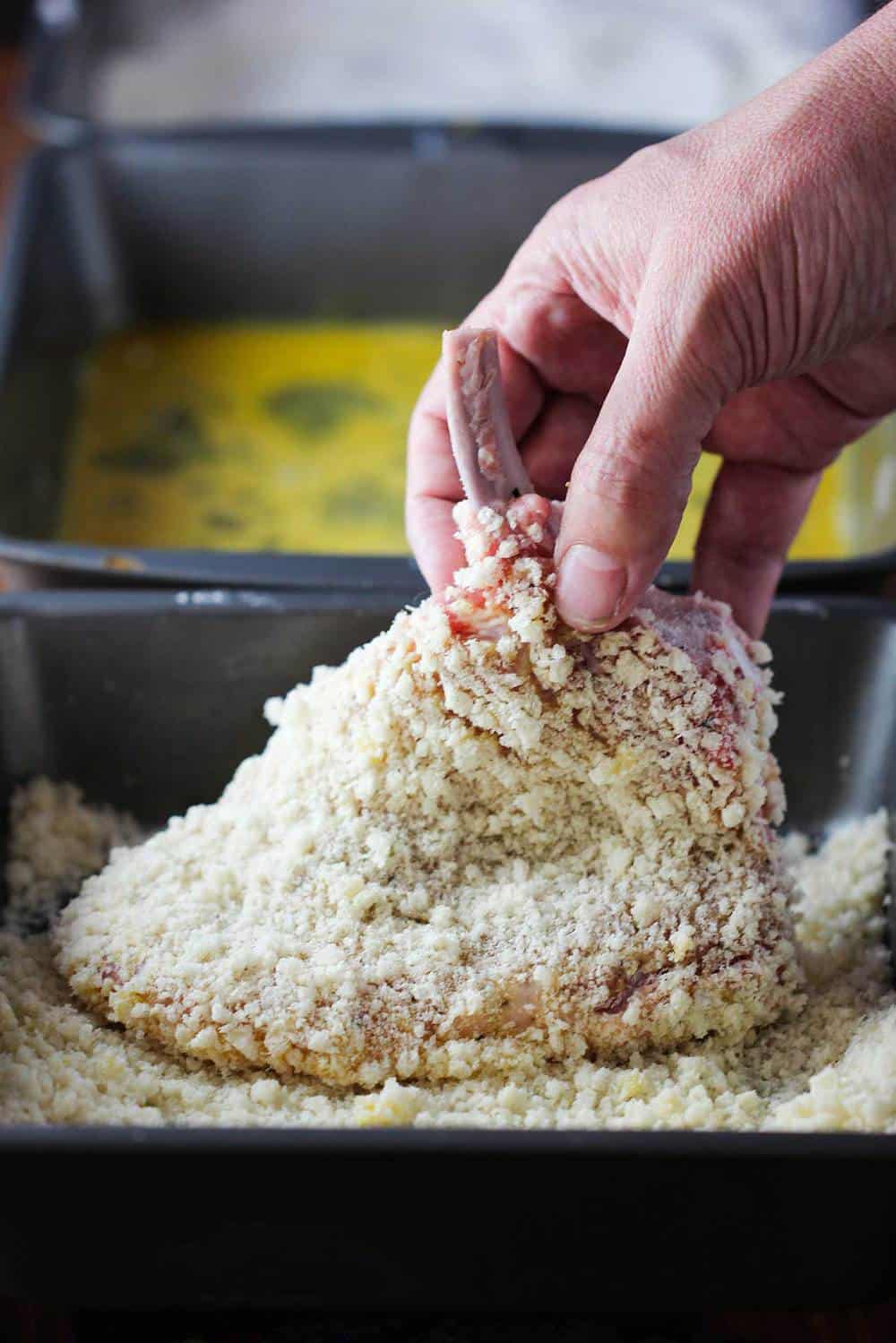 A hand dredging a pork chop in bread crumbs. 