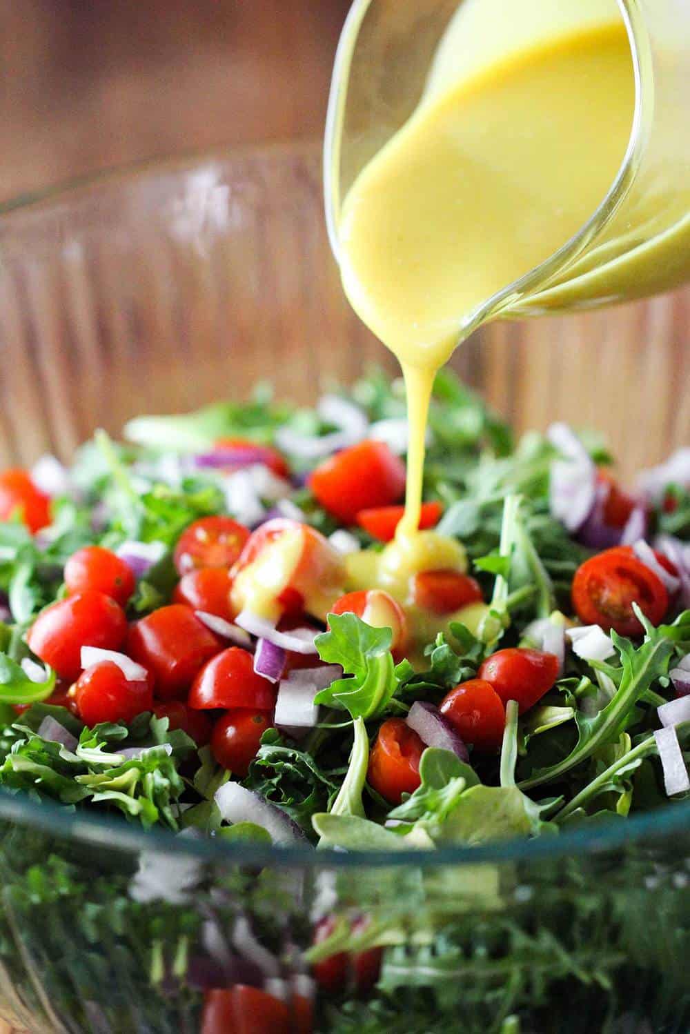 Arugula salad with tomatoes in a glass bowl with vinaigrette being poured over it. 