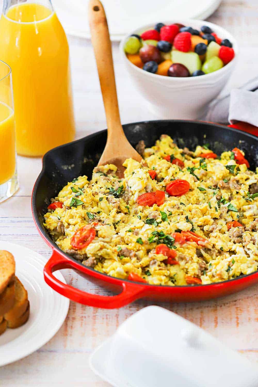 A large red skillet filled with an Italian skillet scramble next to a white bowl filled with fresh fruit.