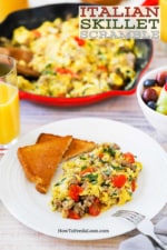 A white dinner plate filled with a large helping of Italian skillet scramble next to a couple pieces of toast cut in half, all next to a large skillet filled with the scrambled dish.