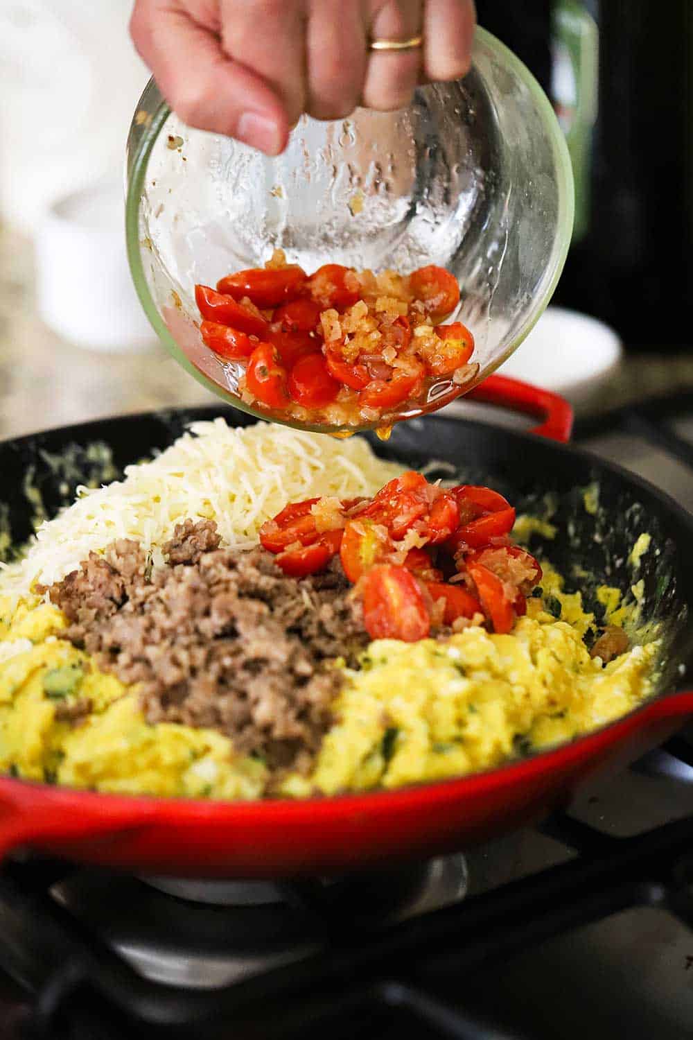A hand transferring sautéd tomatoes from a small glass bowl into a large skillet filled with scrambled eggs, cooked sausage, and shredded mozzarella cheese. 