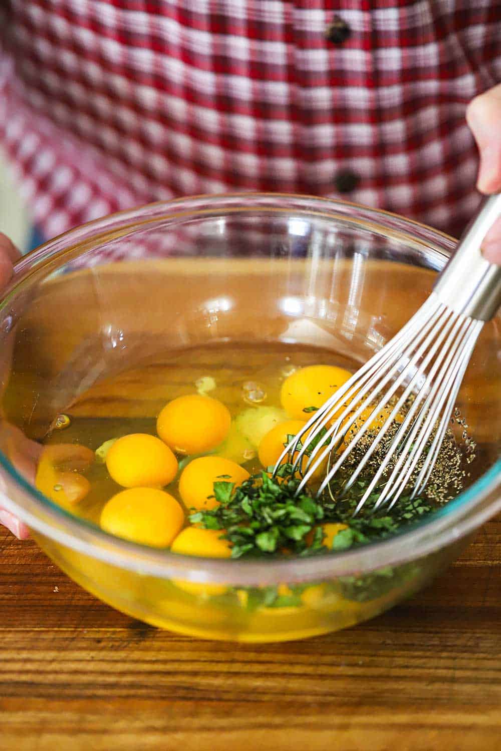 A person using a whisk to lightly beat 12 whole eggs in a glass bowl with chopped basil added in, as well. 