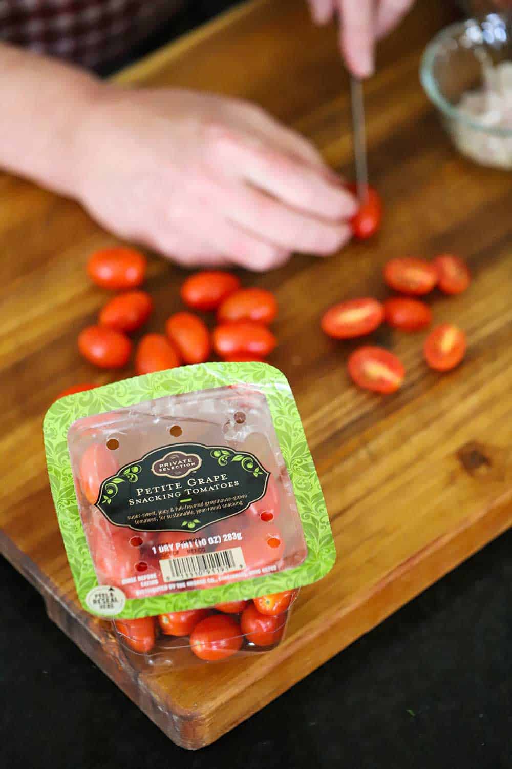 A person using a small knife to slice grape tomatoes in half on a cutting board in front of a package of the tomatoes. 