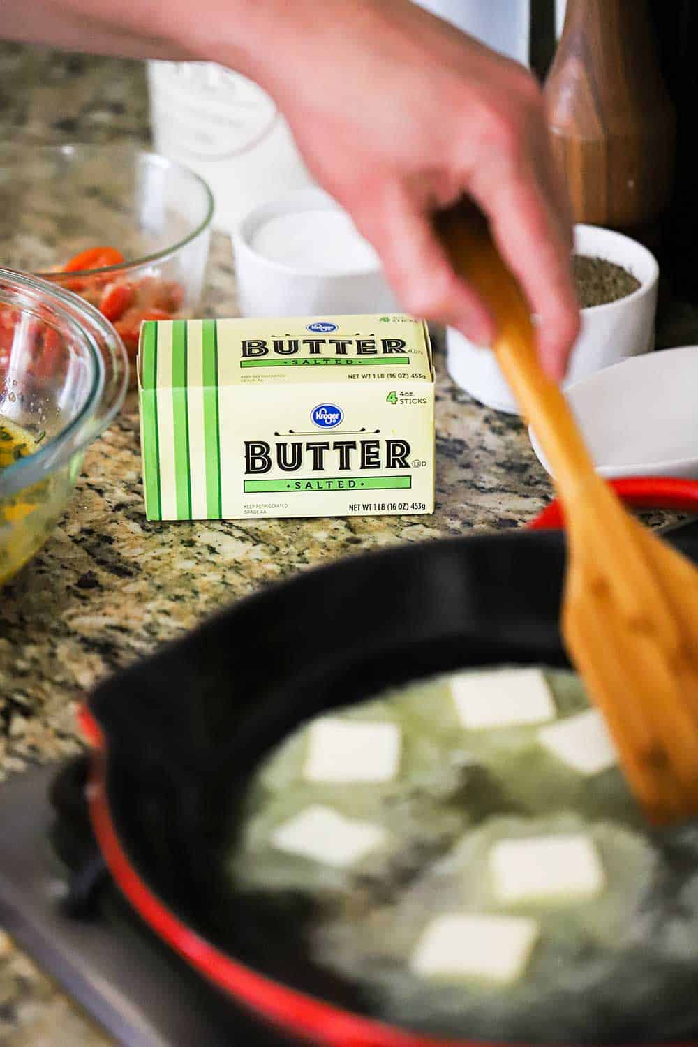 A person using a wooden spatula to help butter melt in a heated skillet all sitting in front of a box of Kroger salted butter. 