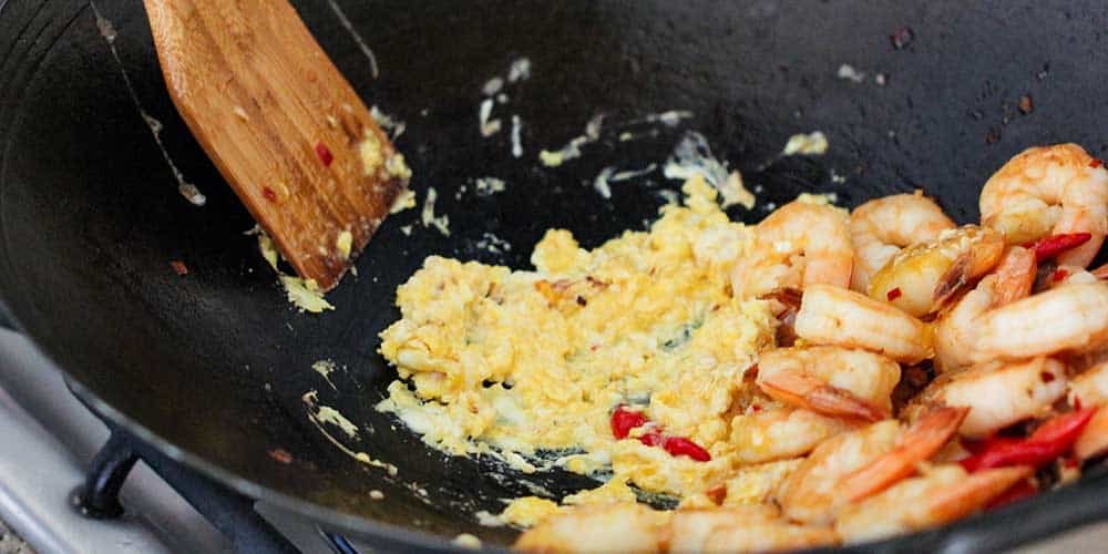 Two eggs being scramble with shrimp and red peppers in a large wok with a wooden spoon. 