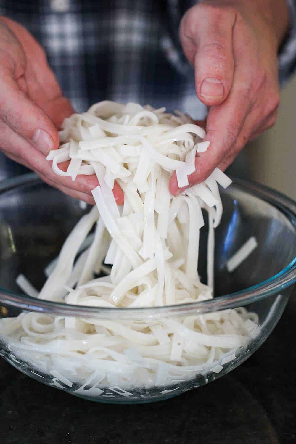 Two hands holding cooked rice noodles in a glass bowl. 