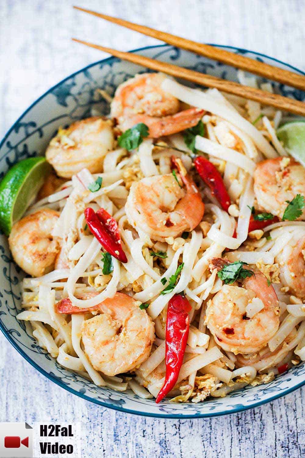 An overhead view of a colorful bowl filled with shrimp pad Thai with two chop sticks on the side of the bowl. 