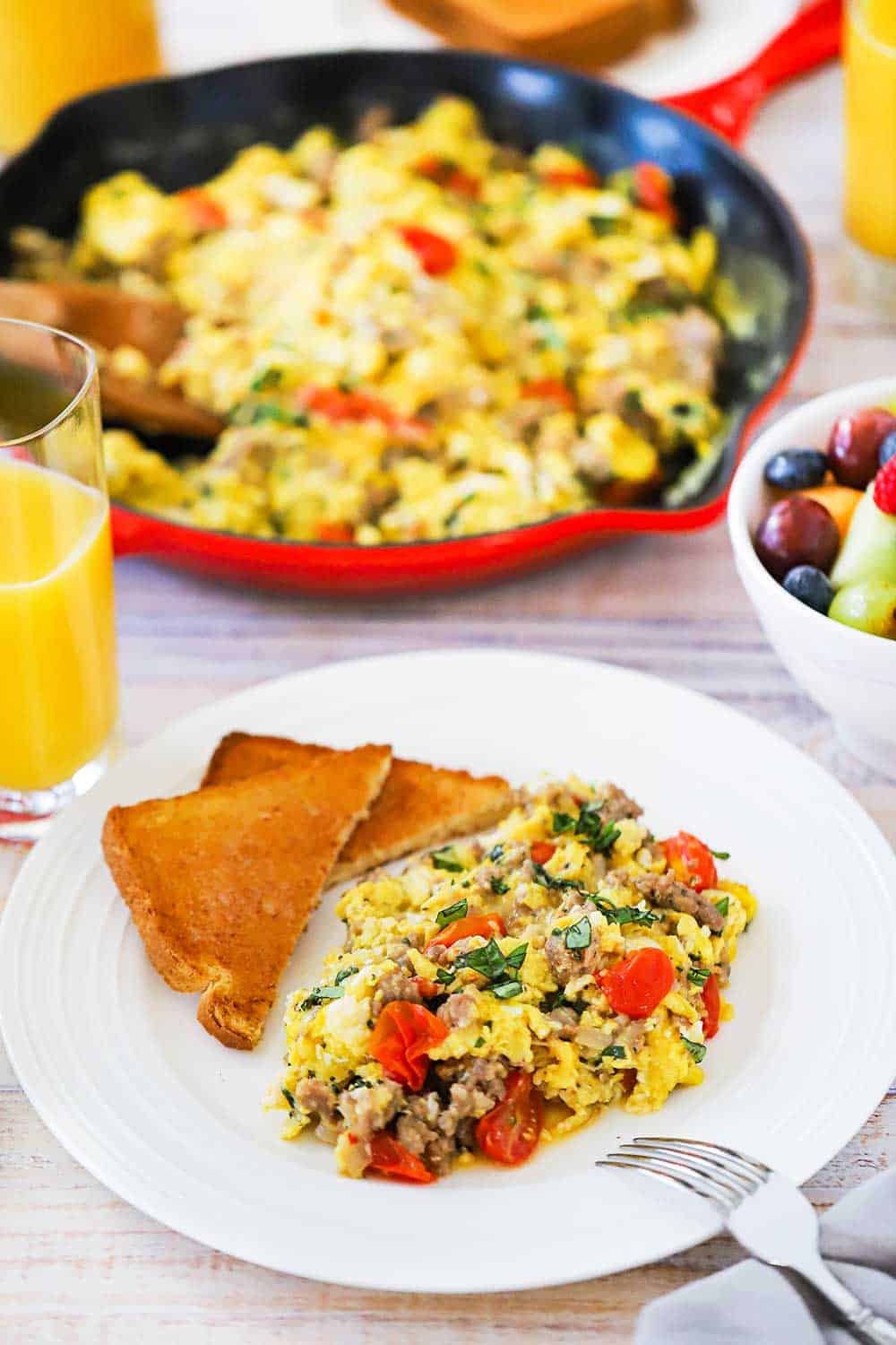 A white dinner plate filled with a large helping of Italian skillet scramble next to a couple pieces of toast cut in half, all next to a large skillet filled with the scrambled dish. 