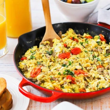 A large red skillet filed with Italian Skillet Scramble with a wooden spoon inserted into it all next to a couple of glasses of orange juice.