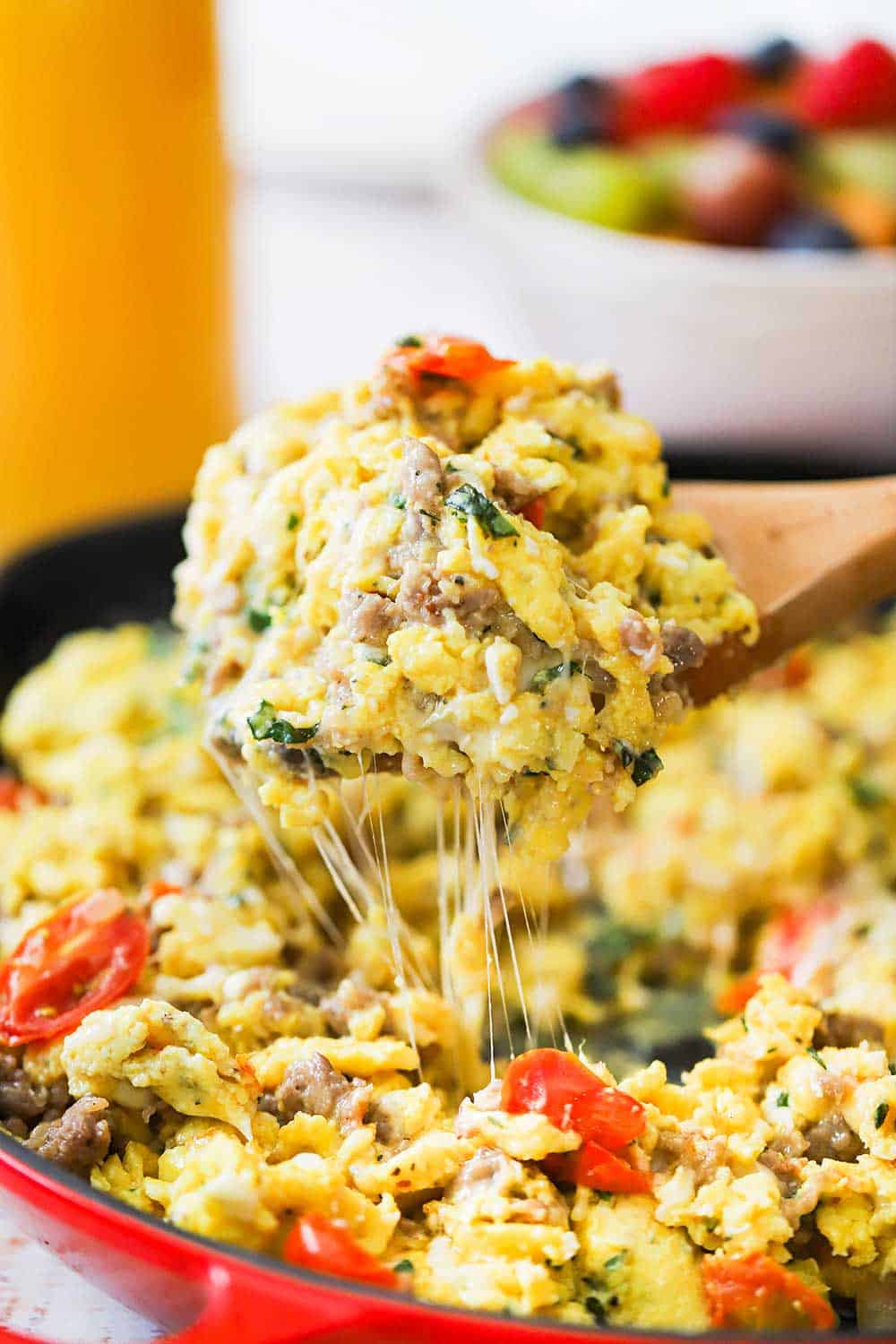 A large wooden spoon lifting up a big helping a cheesy Italian skillet scramble out of a large red skillet. 