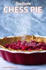 A chess pie in a red pie dish topped with fresh raspberries being sprinkled with powdered sugar from above.