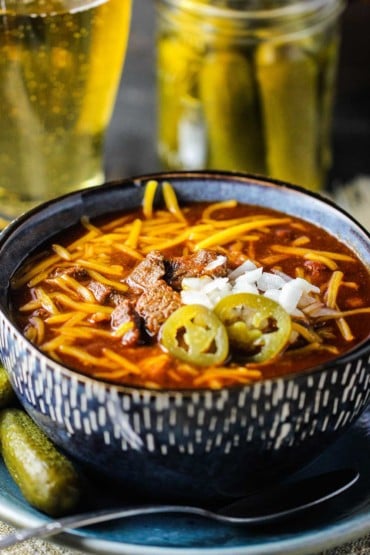 A large blue bowl filled with Texas chili topped with cheese, onions, and jalapenos next to a glass of beer and a jar of pickles.