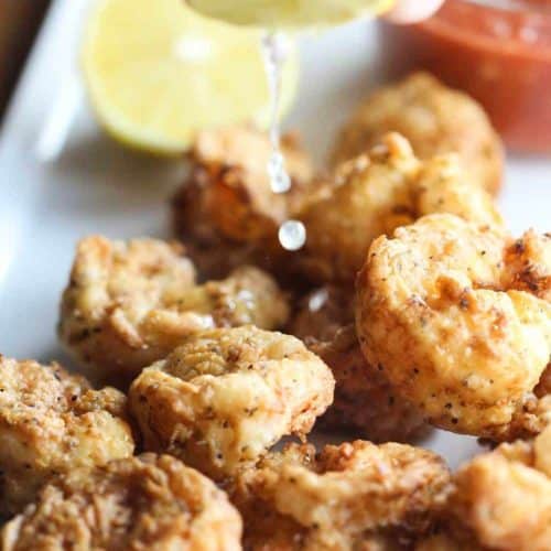 A hand squeezing lemons over Southern Fried Shrimp on a white plate with a bowl of cocktail sauce