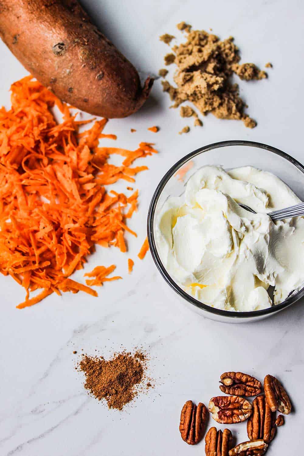 Shredded sweet potatoes next to a whole sweet potato and bowl of cream cheese and pecans. 