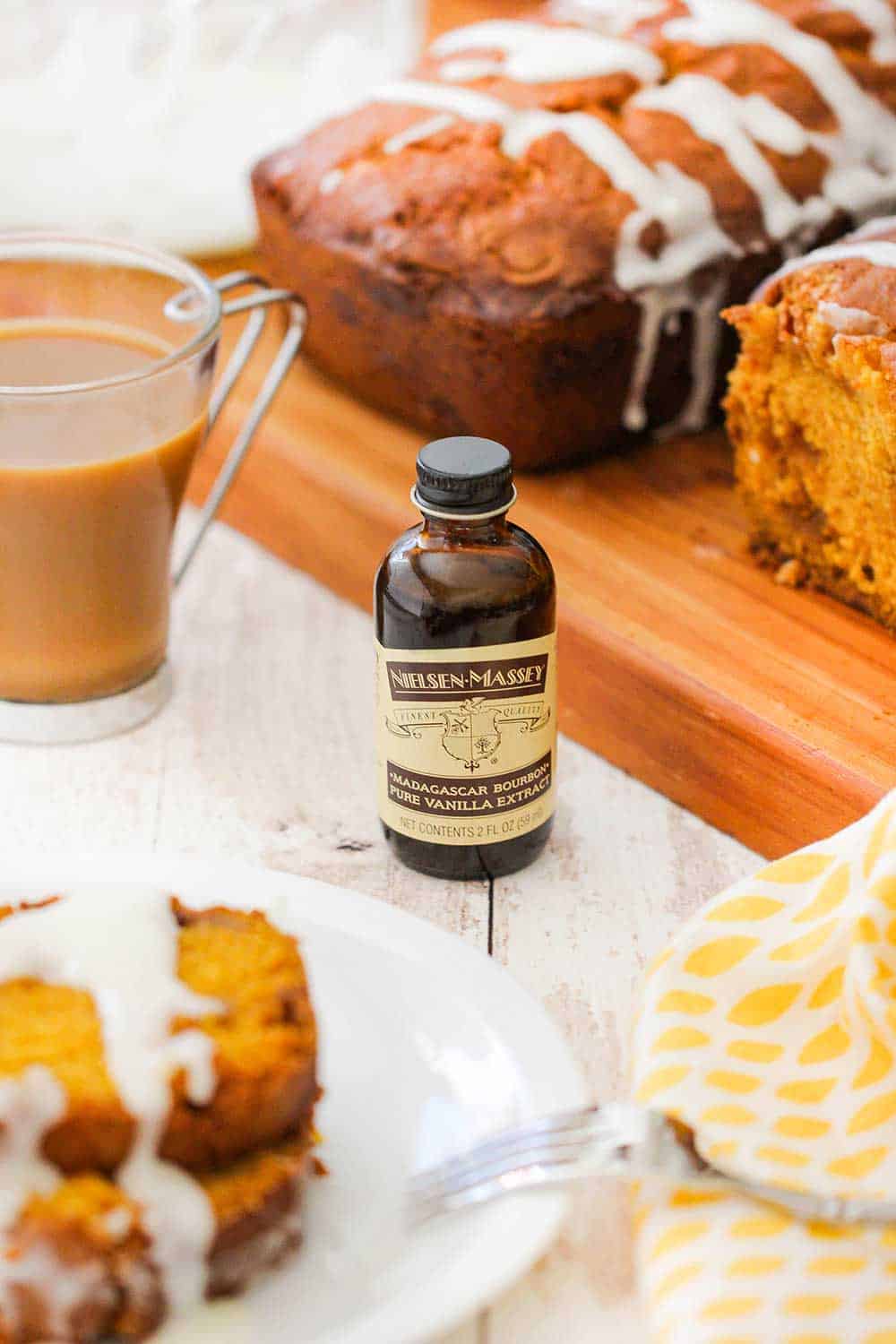 A bottle of vanilla extract between a plate of stacked slices of pumpkin bread and a cutting board with a loaves of bread. 