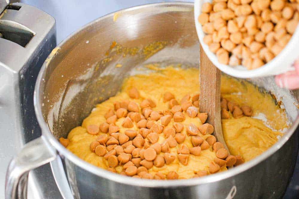 A bowl of butterscotch morsels being mixed into pumpkin bread batter in a metal bowl with a wooden spoon. 