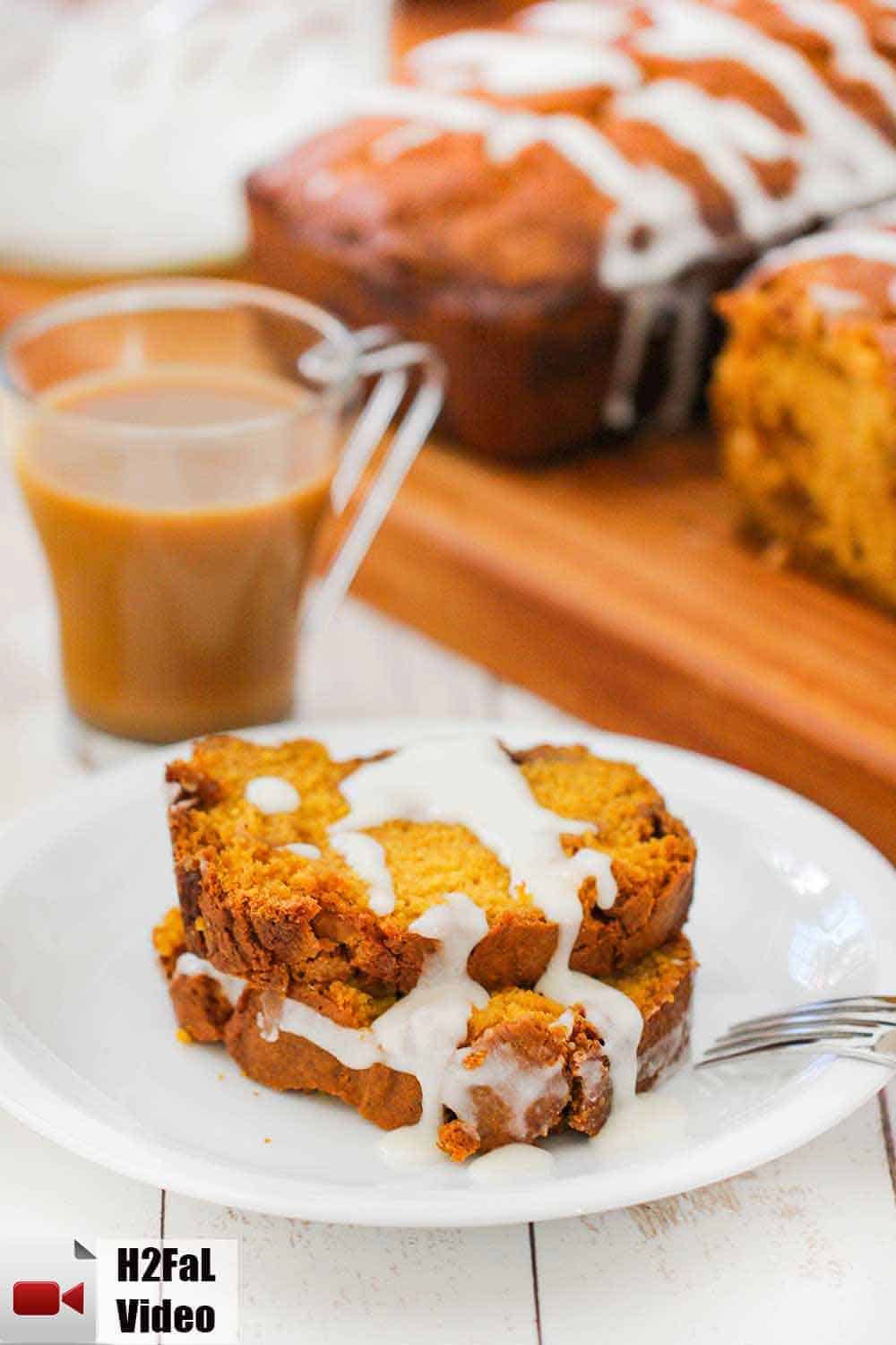 Two slices of pumpkin butterscotch bread on a white plate next to a cup of coffee and two loaves of bread. 