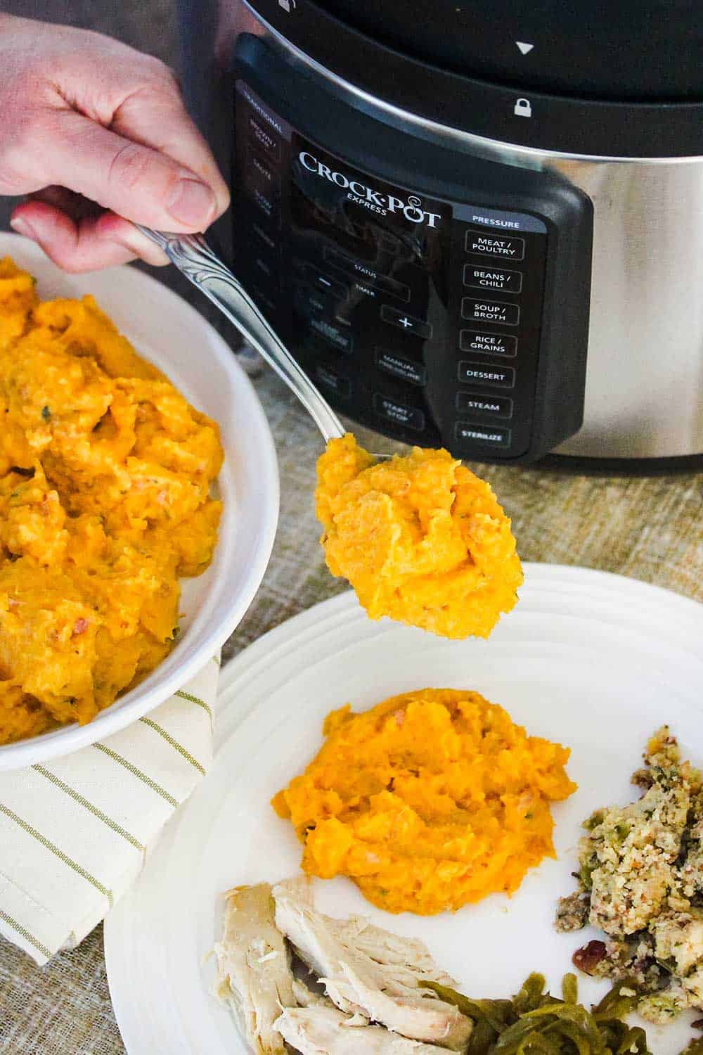 A hand holding a large spoon of mashed butternut squash over a plate of turkey and dressing. 