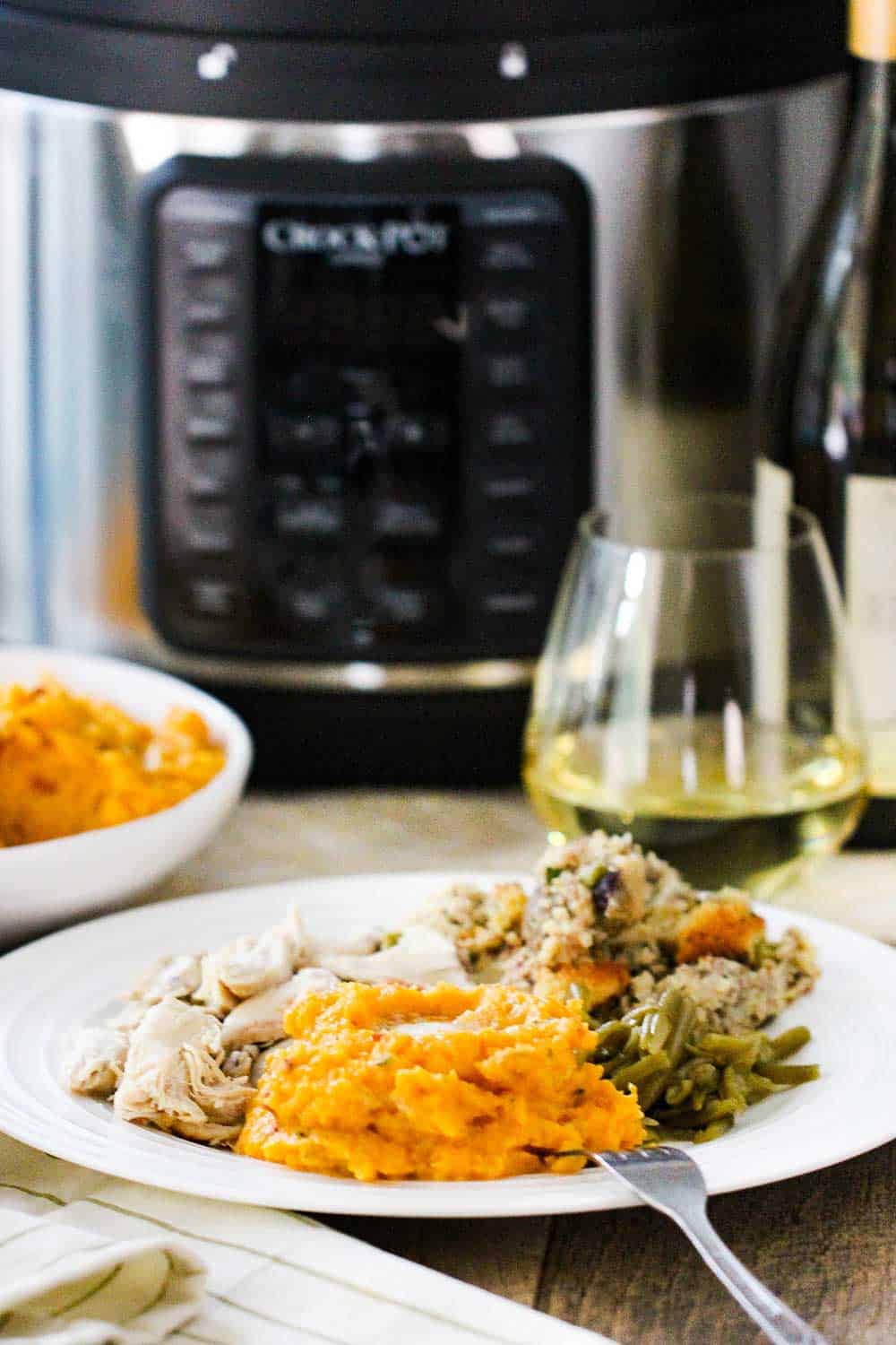 A plate of roasted turkey, mashed butternut squash, dressing and green beans with a glass of white wine near by and a large pressure cooker. 