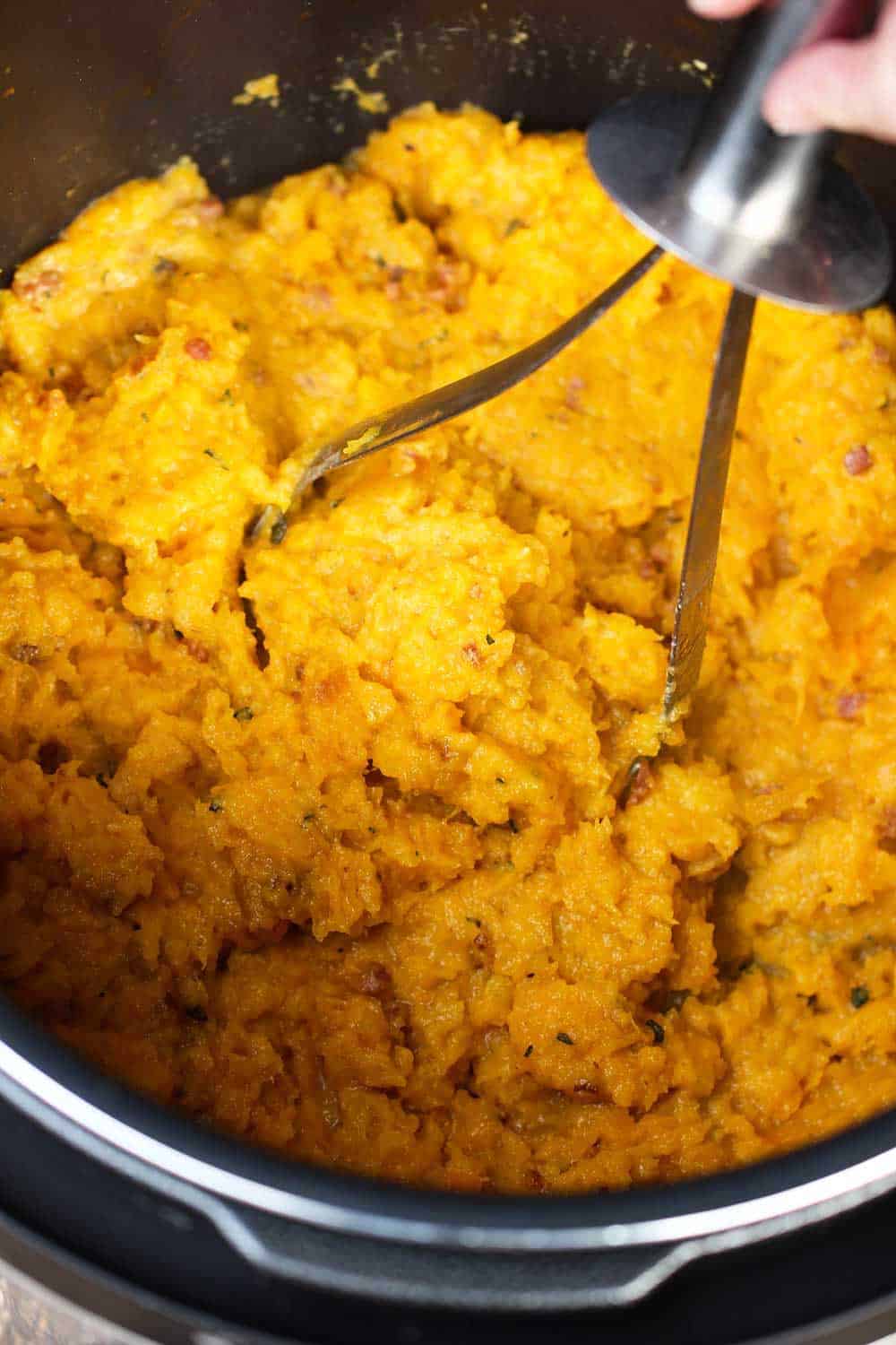 A hand using a potato masher to mash steamed butternut squash. 