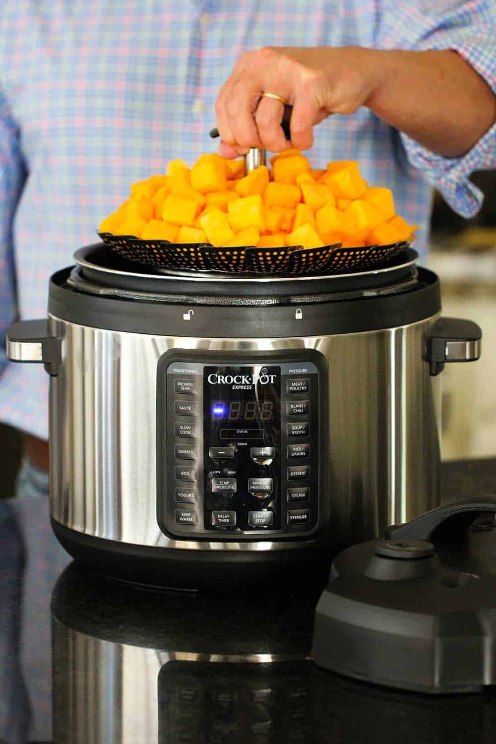 A hand lowering a steamer basket holding cut butternut squash into a 10 qt. pressure cooker. 