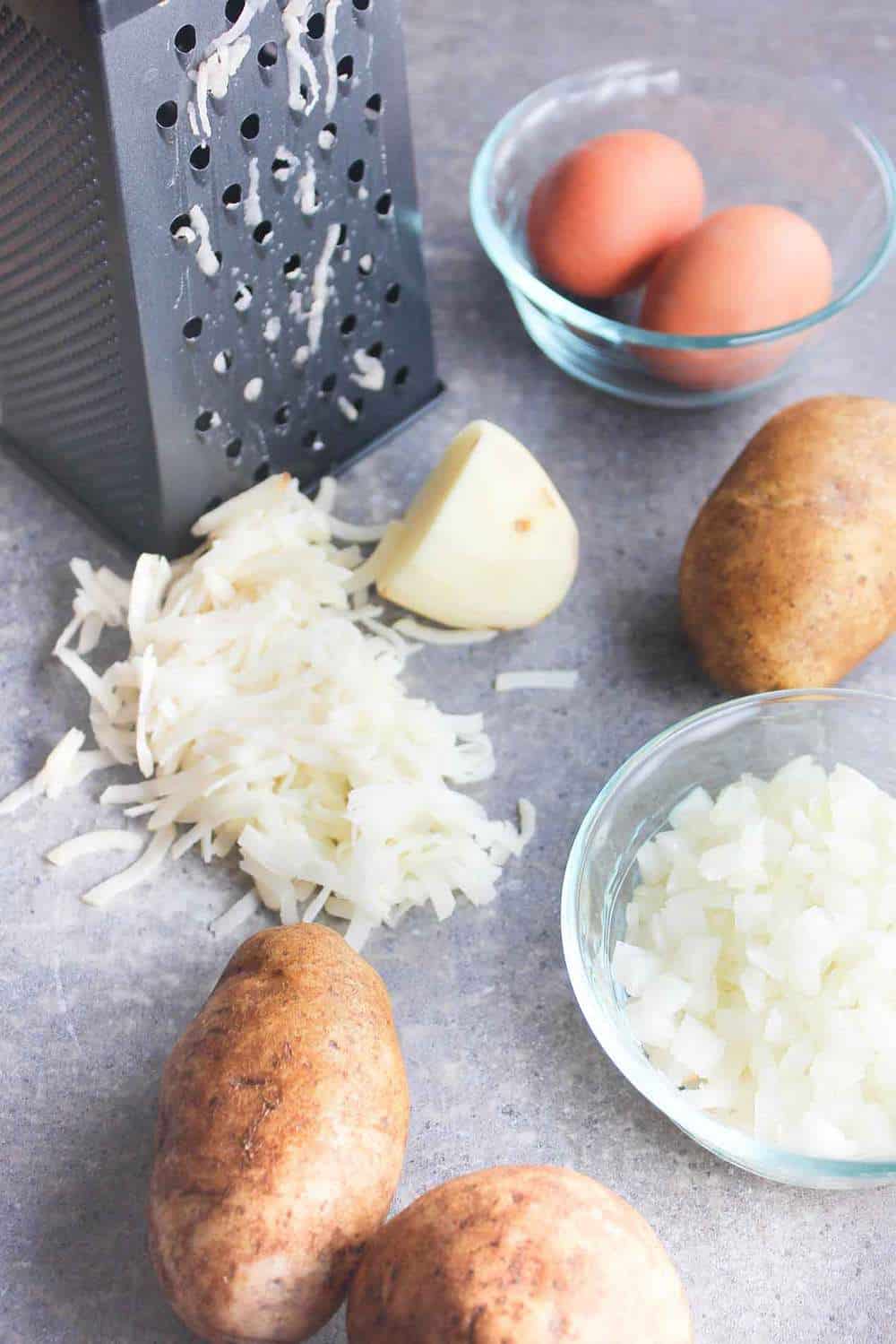 A box grater with grated onion near it sitting next a small bowl filled with two eggs, a bowl of shredded onions, and several russet potatoes