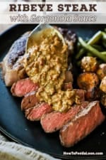 A person using a silver spoon to drizzle a Gorgonzola sauce over a sliced ribeye steak on a black plate also holding roasted potatoes and green beans.