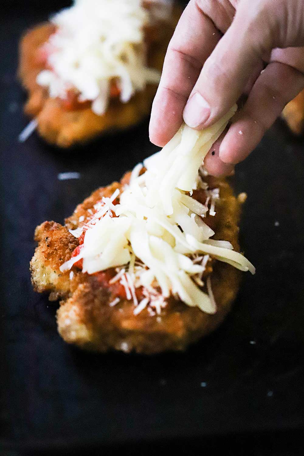 A person placing shredded mozzarella cheese on top of a breaded chicken cutlet that has also been topped with marinara sauce and shredded Parmesan cheese. 