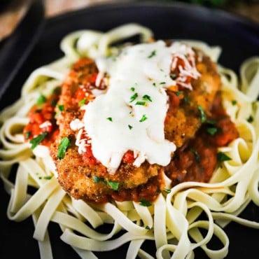 A black dinner plate filled with a pile of cooked fettuccine topped with a chicken parmesan cutlet topped with melted mozzarella cheese.