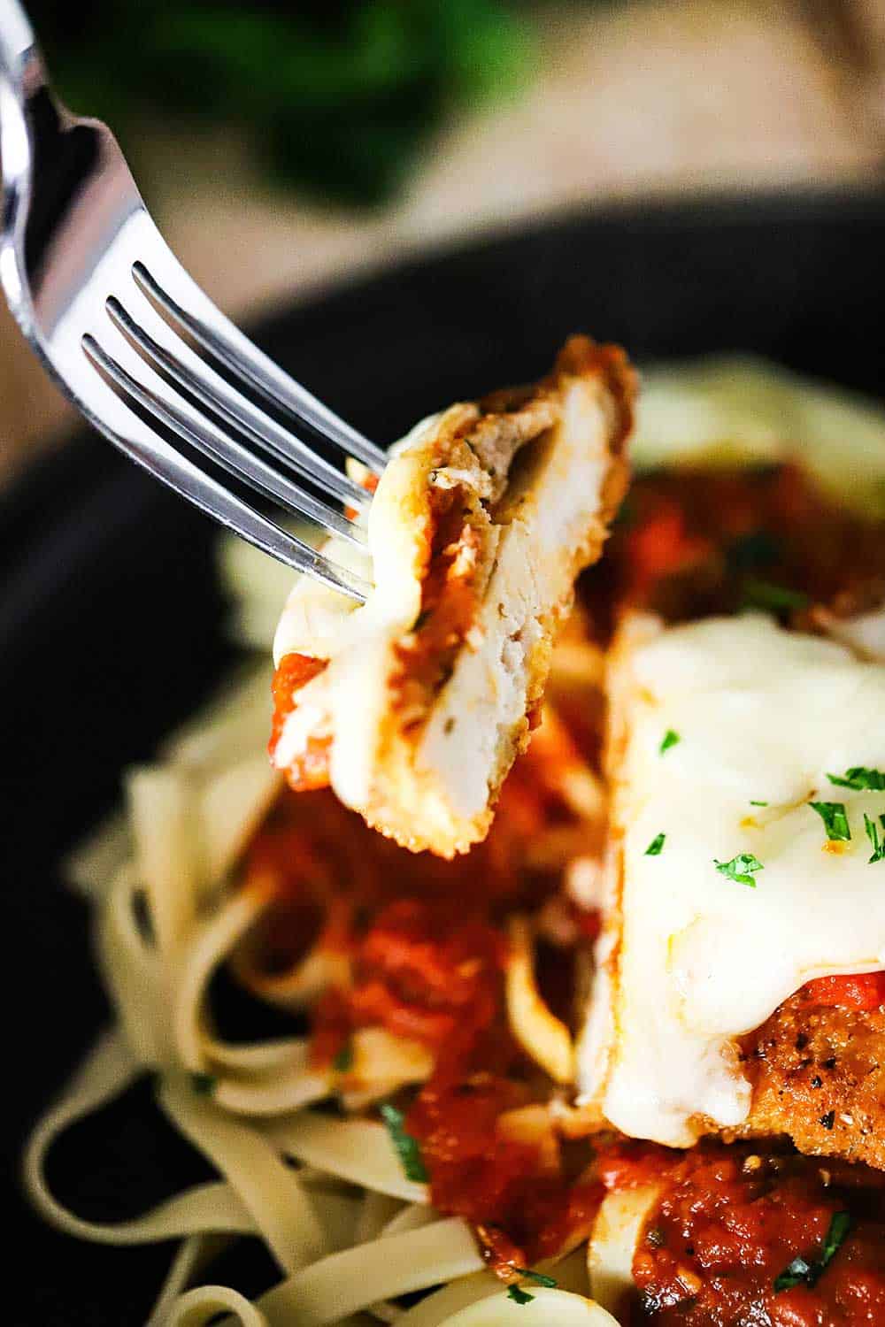 A fork being held up with a slice cut from a chicken parmesan cutlet sitting on a bed of pasta with marinara sauce on it. 