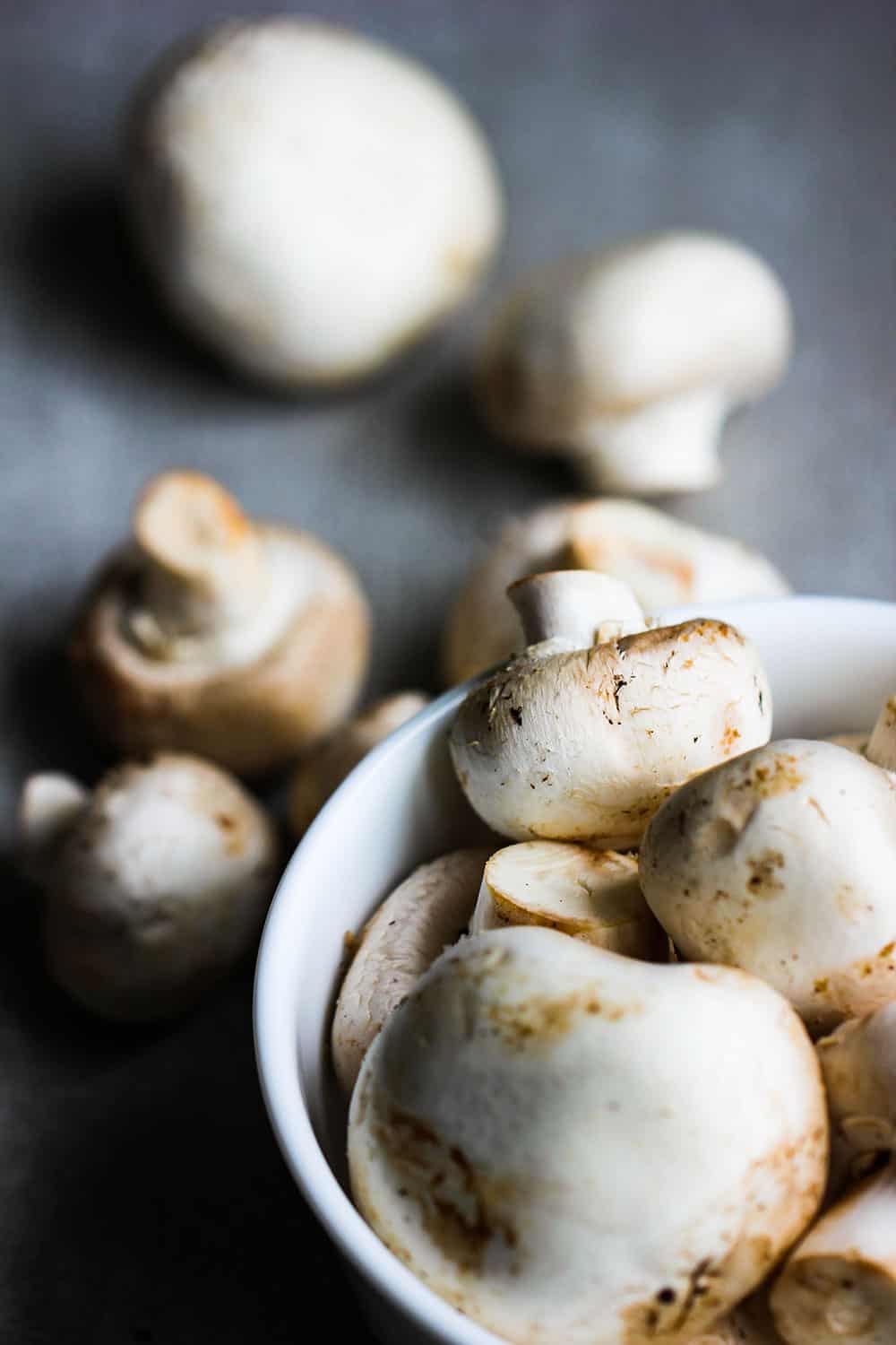 A white bowl holding white button mushrooms with a few extra next to the bowl. 
