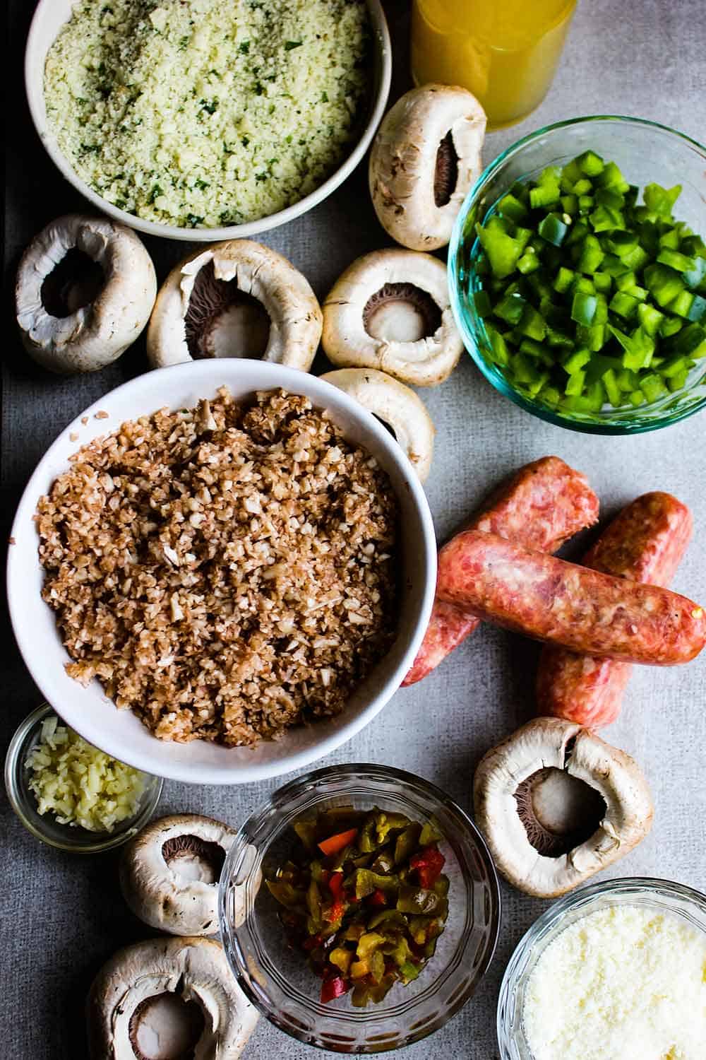 An overhead view of breadcrumbs, chopped mushrooms, links of sausage, and other ingredients for stuffed mushrooms