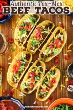Six Tex-Mex Beef Tacos sitting up-right in two taco stands sitting on a cutting board surrounded by bowls of traditional toppings.