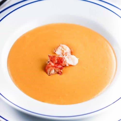 Lobster bisque in a white bowl with blue rim next to a spoon and white napkin