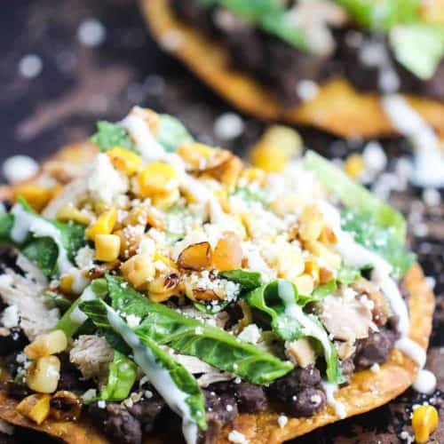 Two black bean and roasted chicken tostadas topped with corn salsa, shredded lettuce, and Mexican crema sitting on a scratched baking sheet.