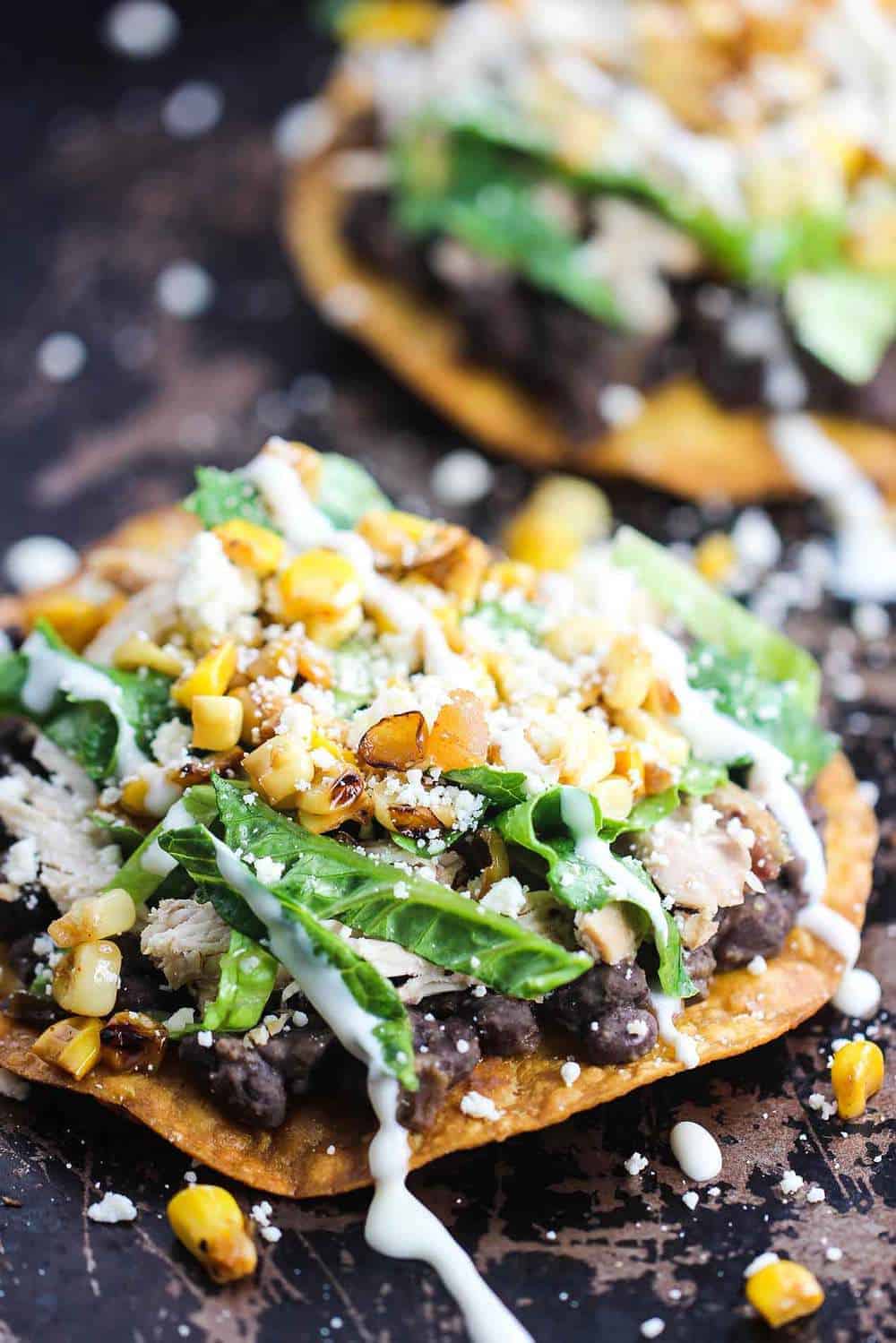 A close-up view of a black bean and roasted chicken tostada that is topped with corn salsa, lettuce, and Mexican crema sitting on a scratchy black baking pan