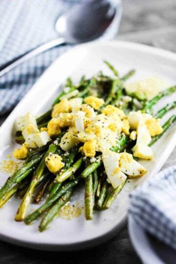 Roasted asparagus, sesame and chopped egg salad on a white dish next to a blue and white checkered napkin with a spoon on top