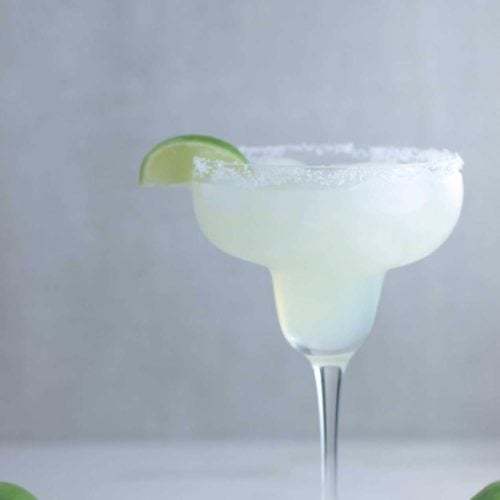 A margarita glass with Mama's Classic Margarita with a slice of lime next to limes