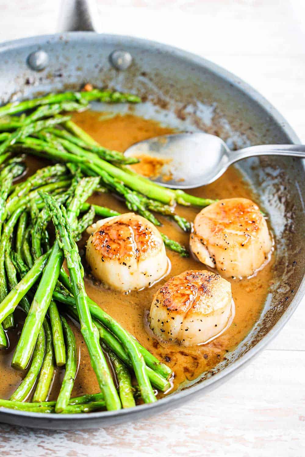 A skillet filled with 3 seared scallops with asparagus in a white wine brown sauce.
