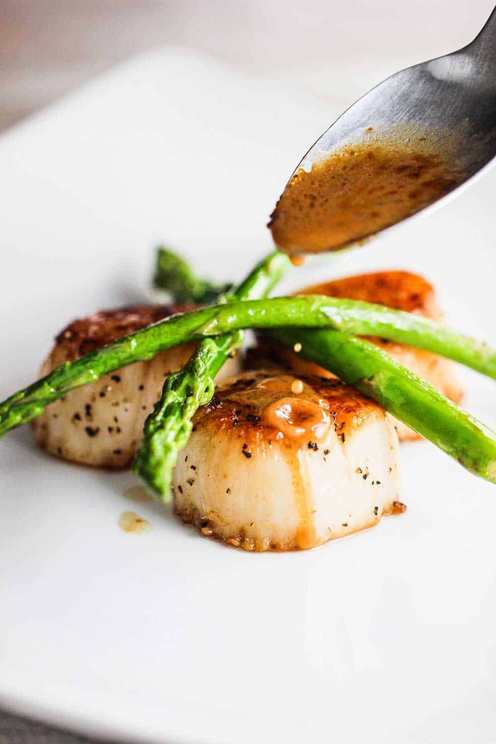 A spoon pouring a brown sauce onto several seared scallops topped with sautéed asparagus.