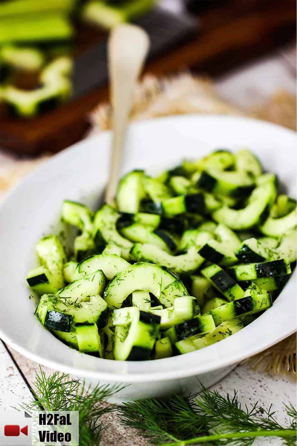 A white bowl of cucumber and dill salad with a gold serving spoon. 