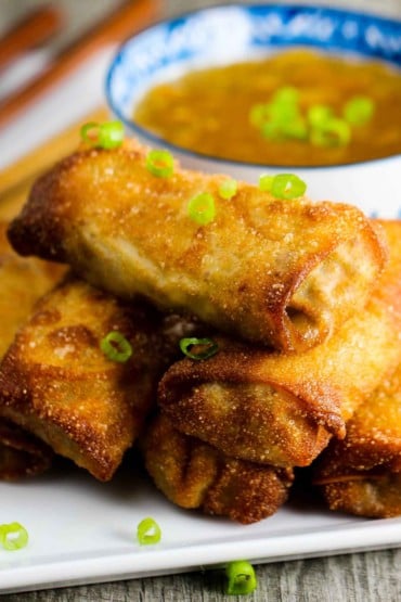 Homemade egg rolls on a white plate next to a patterned bowl with duck sauce