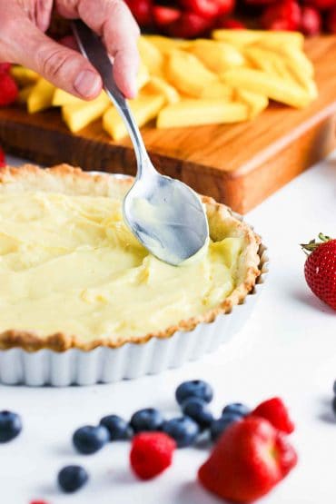 A person using the back of a metal spoon to smooth vanilla custard in a tart pastry shell.