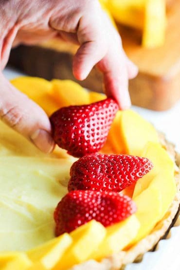 A person adding sliced strawberries on top of custard that is in a tart pastry crust.