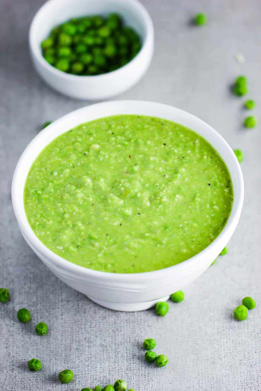 White bowl filled with beautiful pea pesto surround by green peas.