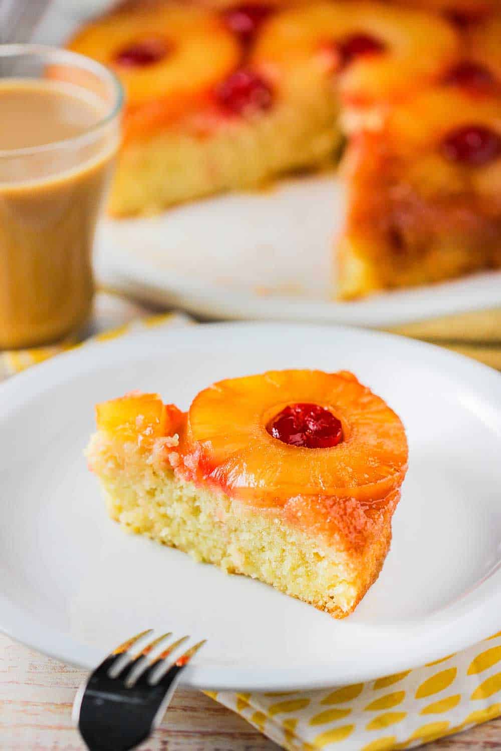 A slice of pineapple upside-down cake on a white plate next to a mug of coffee. 